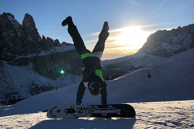 Person doing a handstand on a mountain, with the snowboard in front of them. The sun and other mountains are in the background.