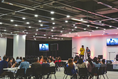 People gathered in groups at tables indoors, while two people on stage are talking into a microphone.